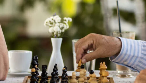 Retirement Community in Boca Raton, FL | Sunscape Boca Raton Mature man enjoying with his wife outdoors and playing chess. Focus is on man's hand holding chess piece.