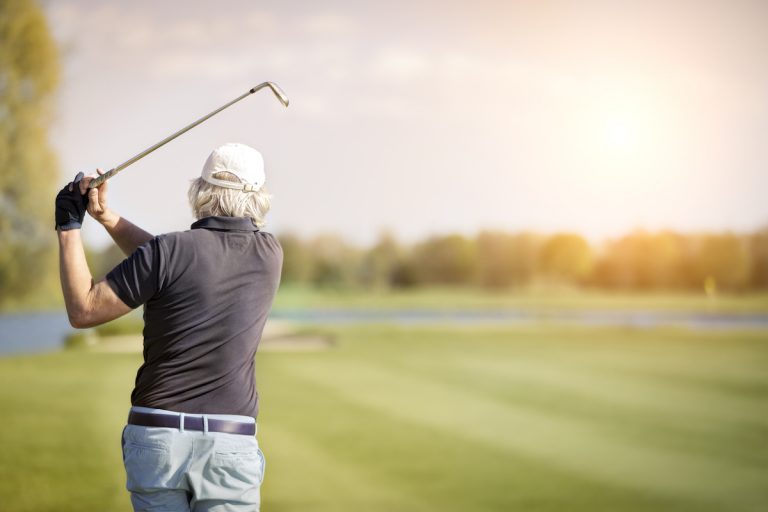 Fore! The Health Benefits of Golf May Surprise You Back of Senior Man Swinging Golf Club at Sunset_Health Benefits of Golf