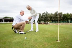 Senior Woman Puts Golf Ball While Husband Bends to Get Closer Look_Retirement Community Boca Raton
