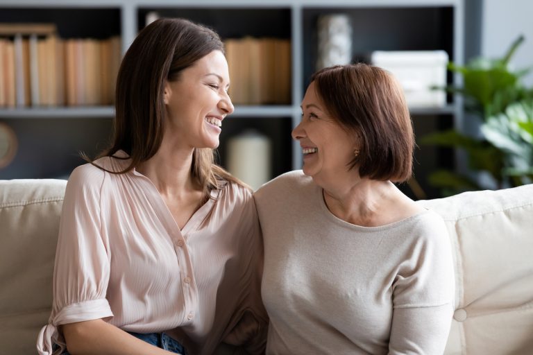 Should Mom Move into Senior Living in Boca Raton? Senior Woman and Adult Daughter Laughing on Couch_Senior Living in Boca Raton