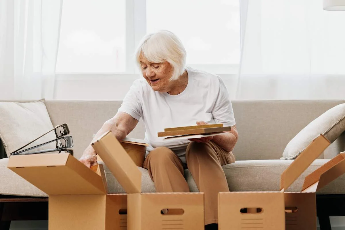elderly woman sits on a sofa at home with boxes. collecting things with memories albums with photos and photo frames moving to a new place cleaning things and a happy smile. Lifestyle retirement.
